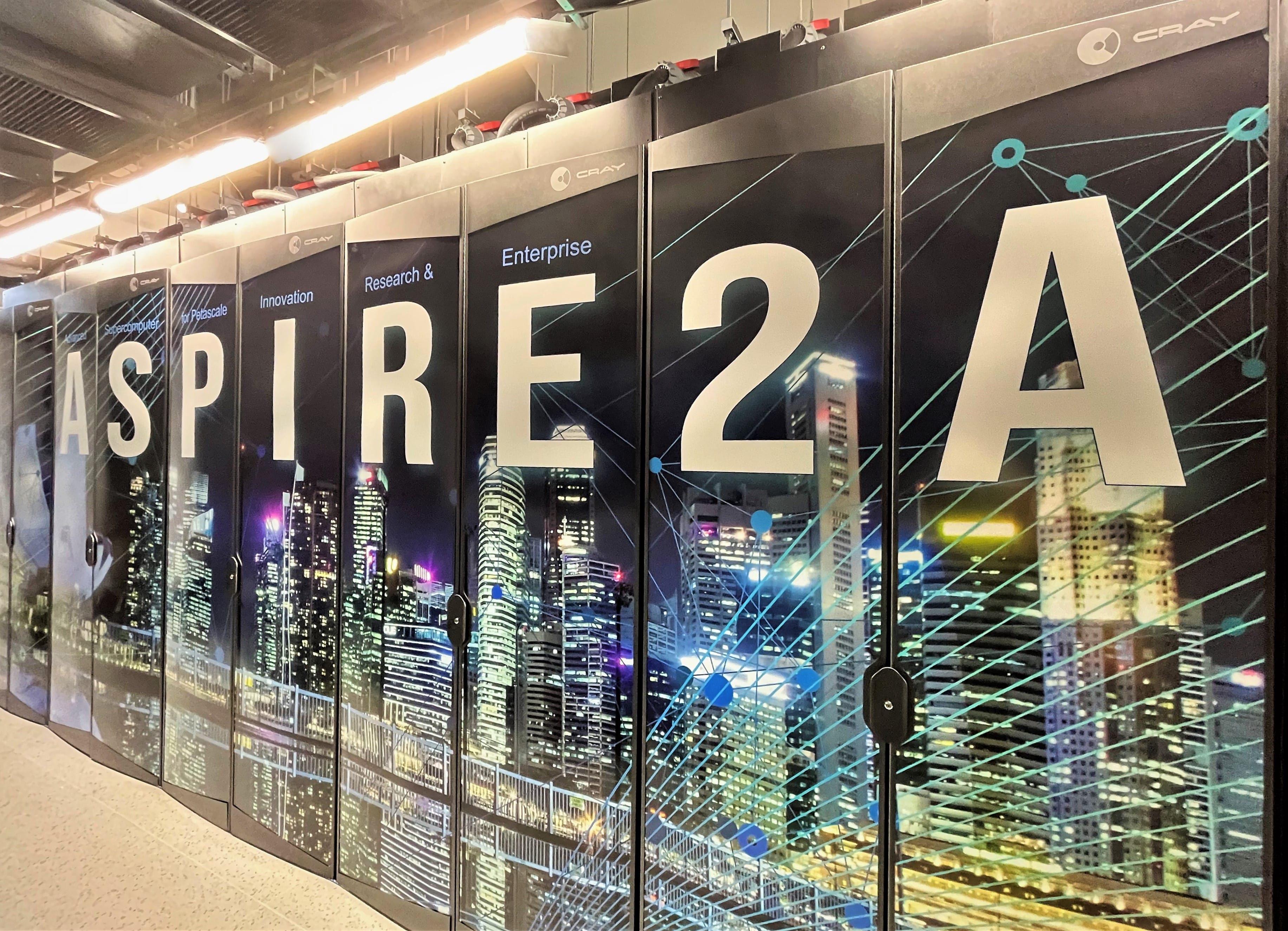 A row of Cray supercomputer cabinets branded ASPIRE 2A with a city skyline graphic in a data center.